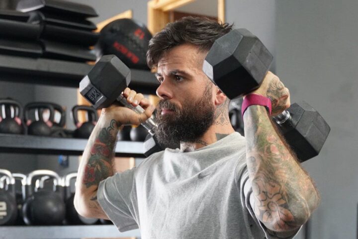 Scott Britton holding dumbbells by his shoulders wearing a pink Whoop tracker on his wrist