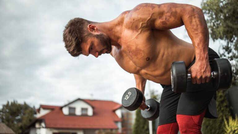 Shirtless muscular man performs bent-over row with dumbbells outside