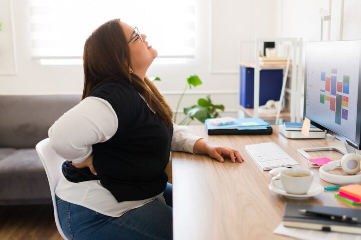 Woman sitting at desk holds her lower back and winces in pain